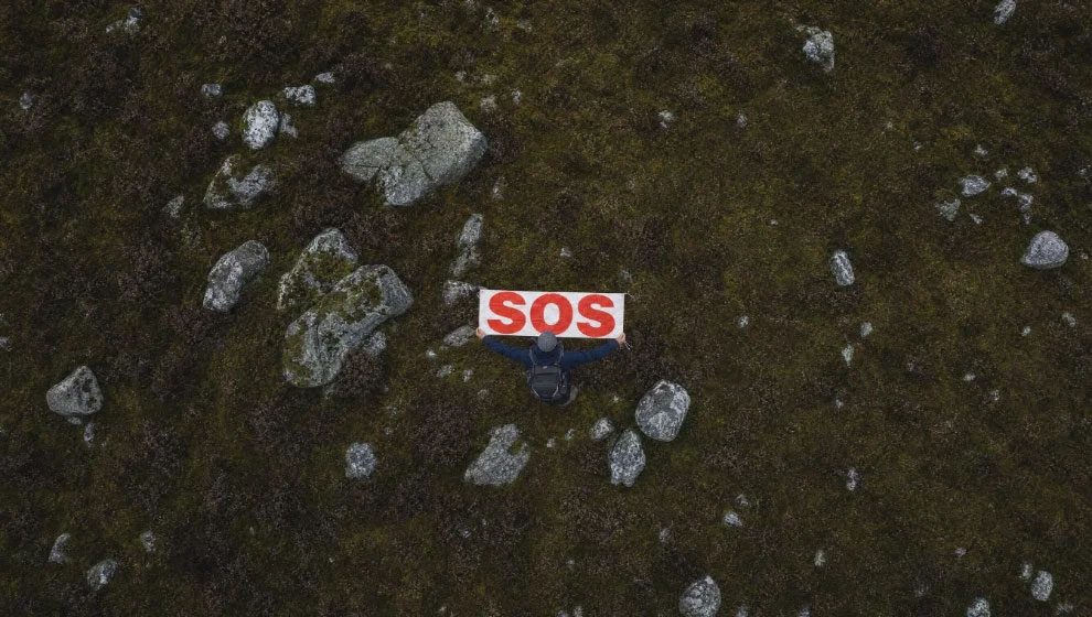 A walker lying on dark moorland holding the WalkSOS banner, photographed from directly
                  above. The ground is olive and brown, scattered with grey rock. The banner is the only
                  straight-edged object in the frame.